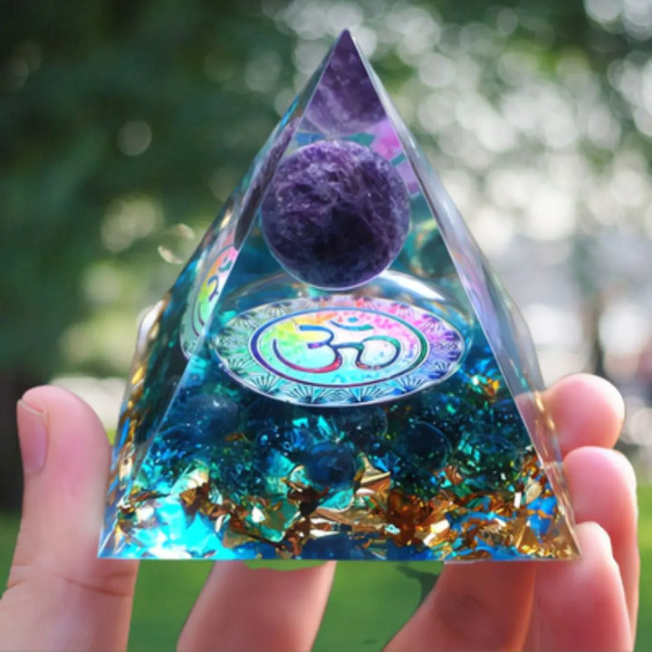 Orgonite Pirâmide com Símbolo Om e Ametista Roxa sendo segurada ao ar livre, com árvores desfocadas ao fundo, destacando a esfera de ametista e o símbolo Om