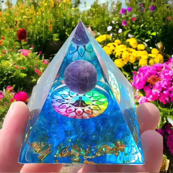 Orgonite Pirâmide 7 Chakras com Ametista Roxa sendo segurada em um jardim florido, destacando suas cores vibrantes e a esfera de ametista