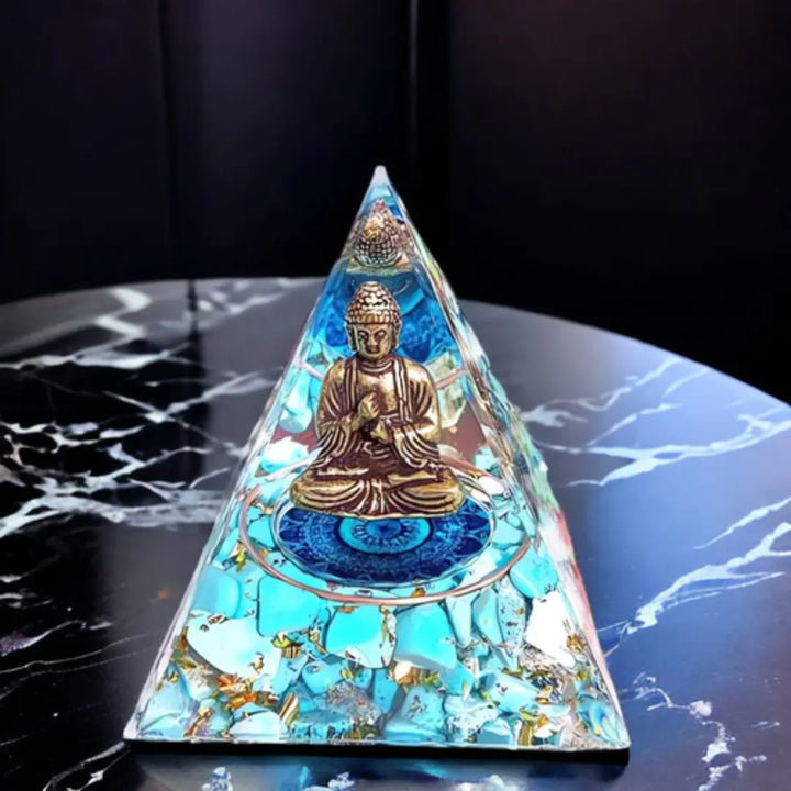 Orgonite Pirâmide Buda Meditação com Quartzo Azul, apresentando uma figura de Buda meditando em meio a fragmentos de quartzo azul brilhante sobre uma mesa de mármore.