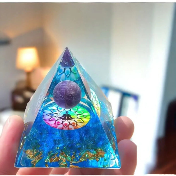 Orgonite Pirâmide 7 Chakras com Ametista Roxa sendo segurada dentro de casa, com uma lâmpada acesa ao fundo, mostrando sua beleza e energia